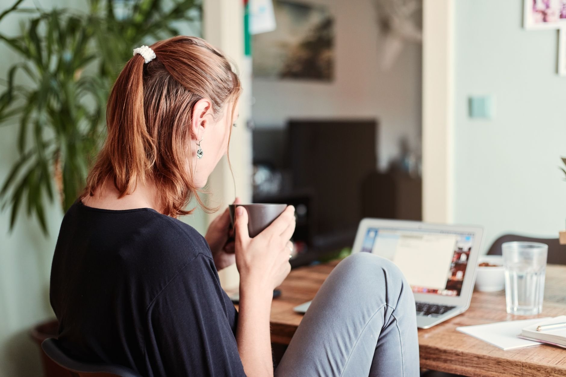 A woman in a cozy home office setting, sipping coffee while working on a laptop.