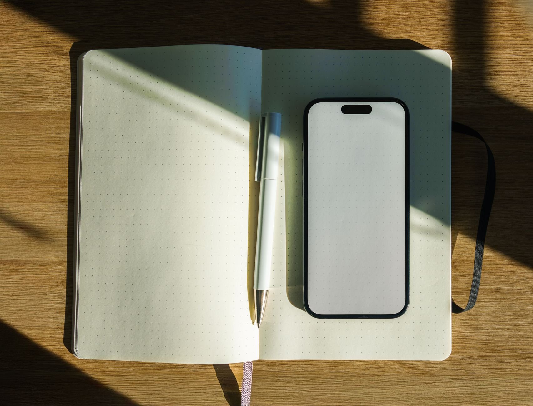 A minimalistic sunlit workspace featuring an open notebook, pen, and smartphone on a wooden desk.